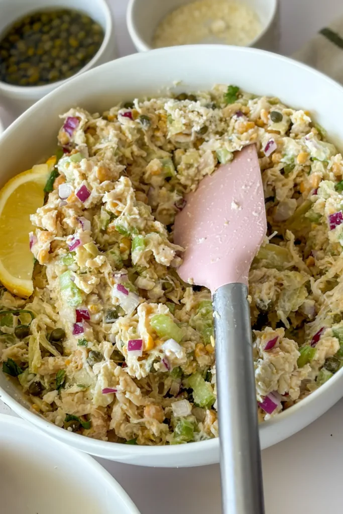 A spatula mixing the chicken salad in a large white bowl on a counter.