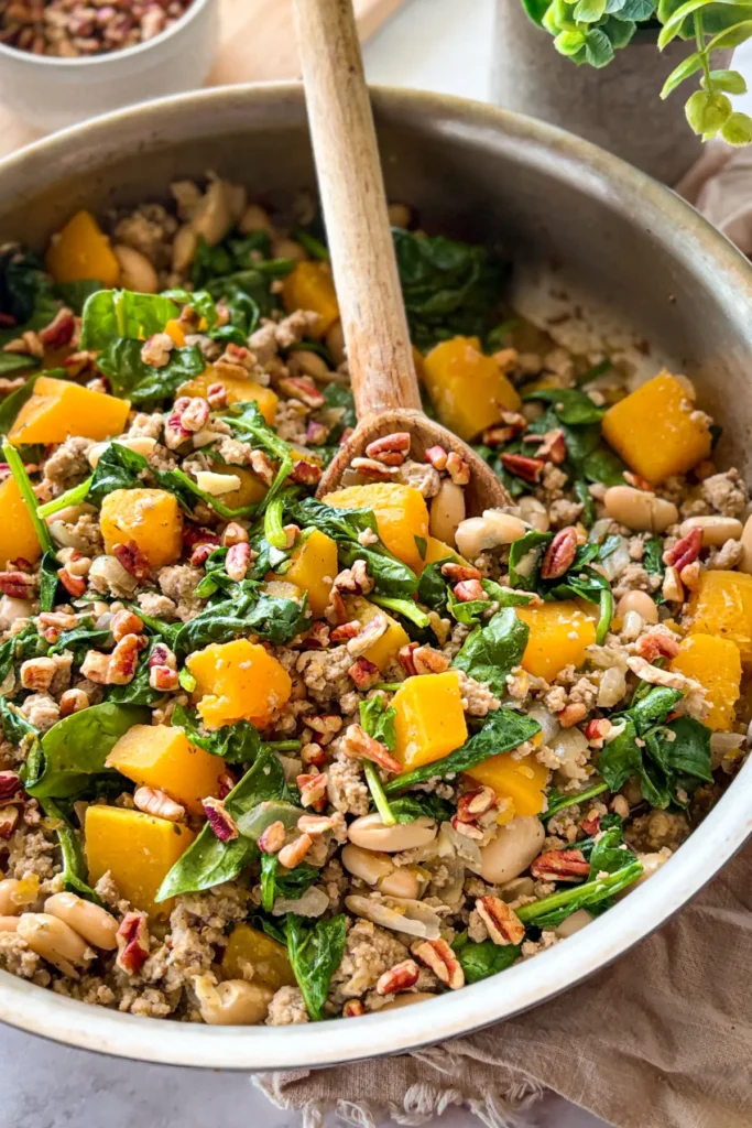 A butternut squash and ground turkey skillet with a wooden spoon in it on a table.