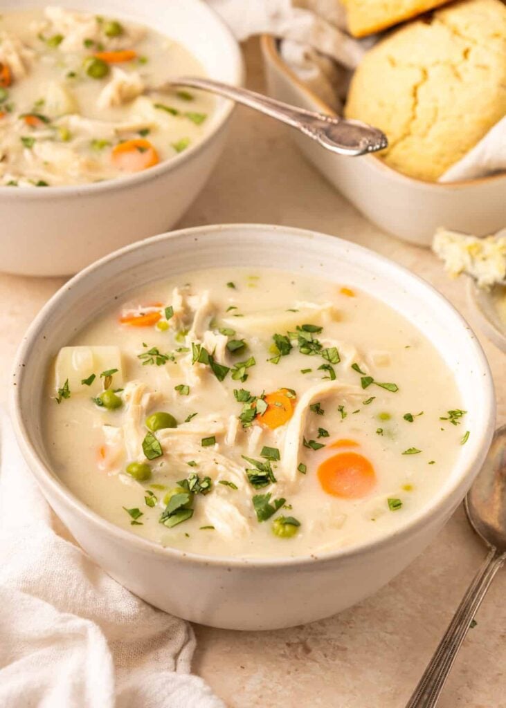 Two bowls of creamy chicken pot pie soup on a table next to a bowl of biscuits.