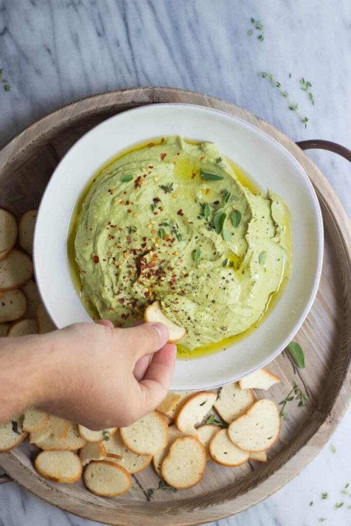 A large bowl of white bean and chickpea dip on a plate with bread crisps.