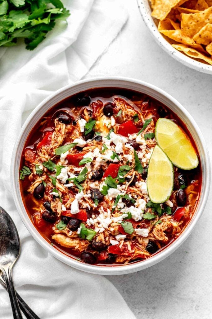 A birds-eye view of a bowl of buffalo chicken chili topped with cheese and lime wedges on a table.