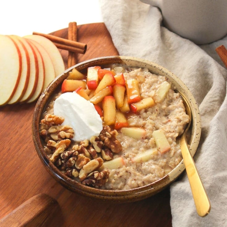 A bowl of oatmeal with warm cinnamon apples, whipped topping and pecans on a wooden cutting board garnished with apple slices.