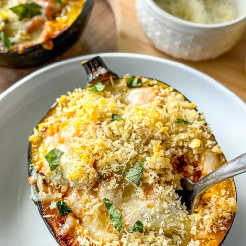 A spoon digging into a roasted stuffed acorn squash on a white plate on a table.