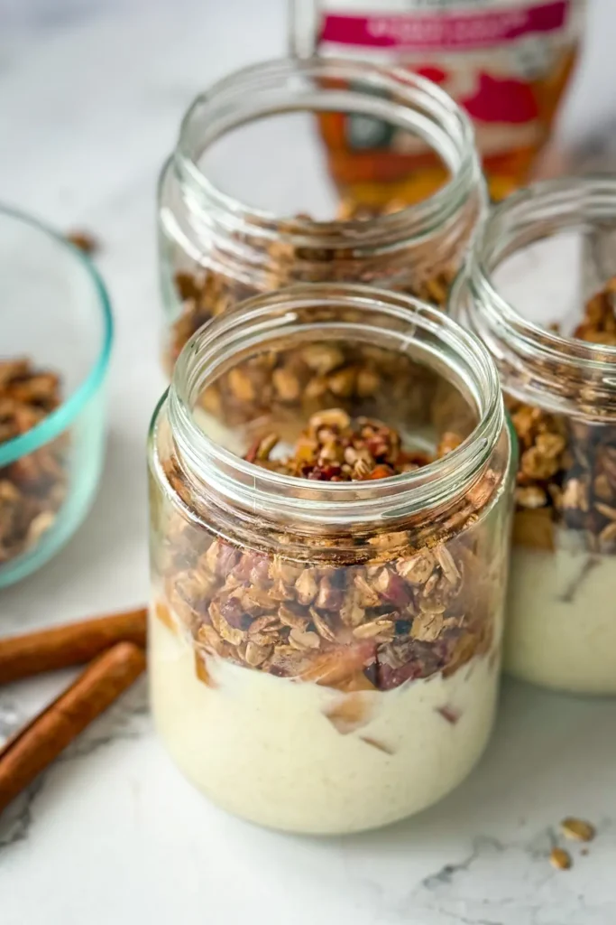 Three yogurt apple crisp jars on a counter next to a bowl of pecans and some maple syrup.