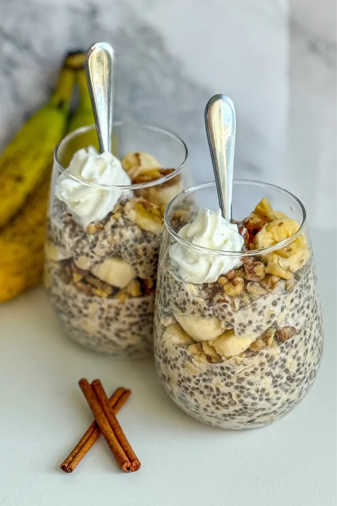 Two glasses filled with banana cinnamon overnight oats topped with whipped cream with spoons in them on a counter.
