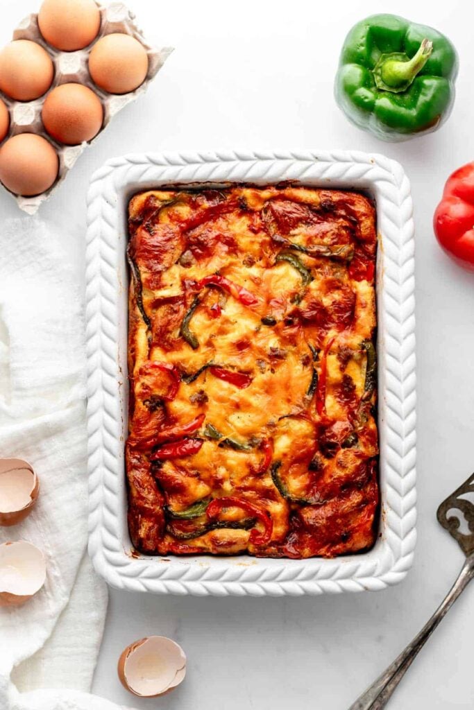 A birds-eye view of a cozy breakfast casserole in a white baking dish on a white counter with garnishes like peppers and eggs on the side.