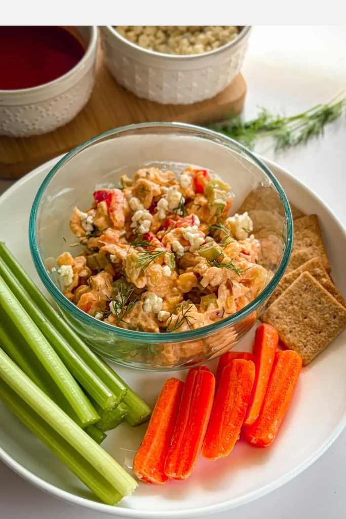 A close-up of a bowl of buffalo chicken snack dip on a plate with celery, carrots and Wheat Thins.