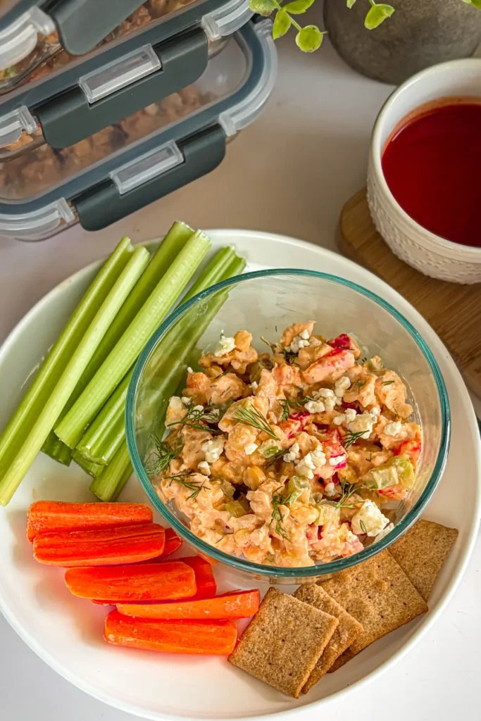A snack plate with buffalo chicken dip, celery, carrots, and Wheat Thins on a counter next to small snack containers of dip.