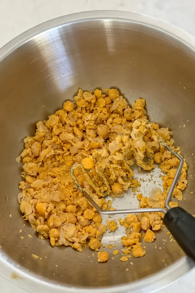 Mashed chickpeas on the bottom of a mixing bowl with a potato masher in it.