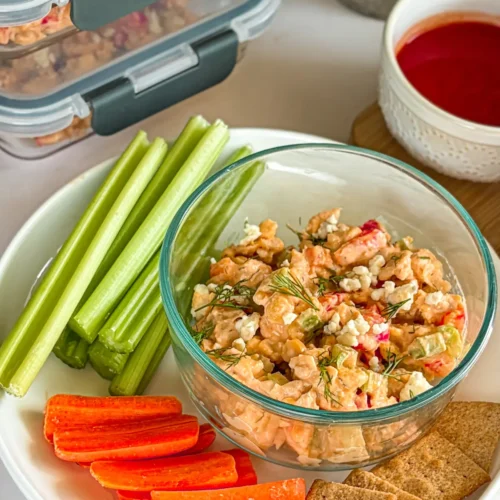 A loaded buffalo chicken dip snack plate with celery, carrots, and crackers on a table next to dip snack containers.