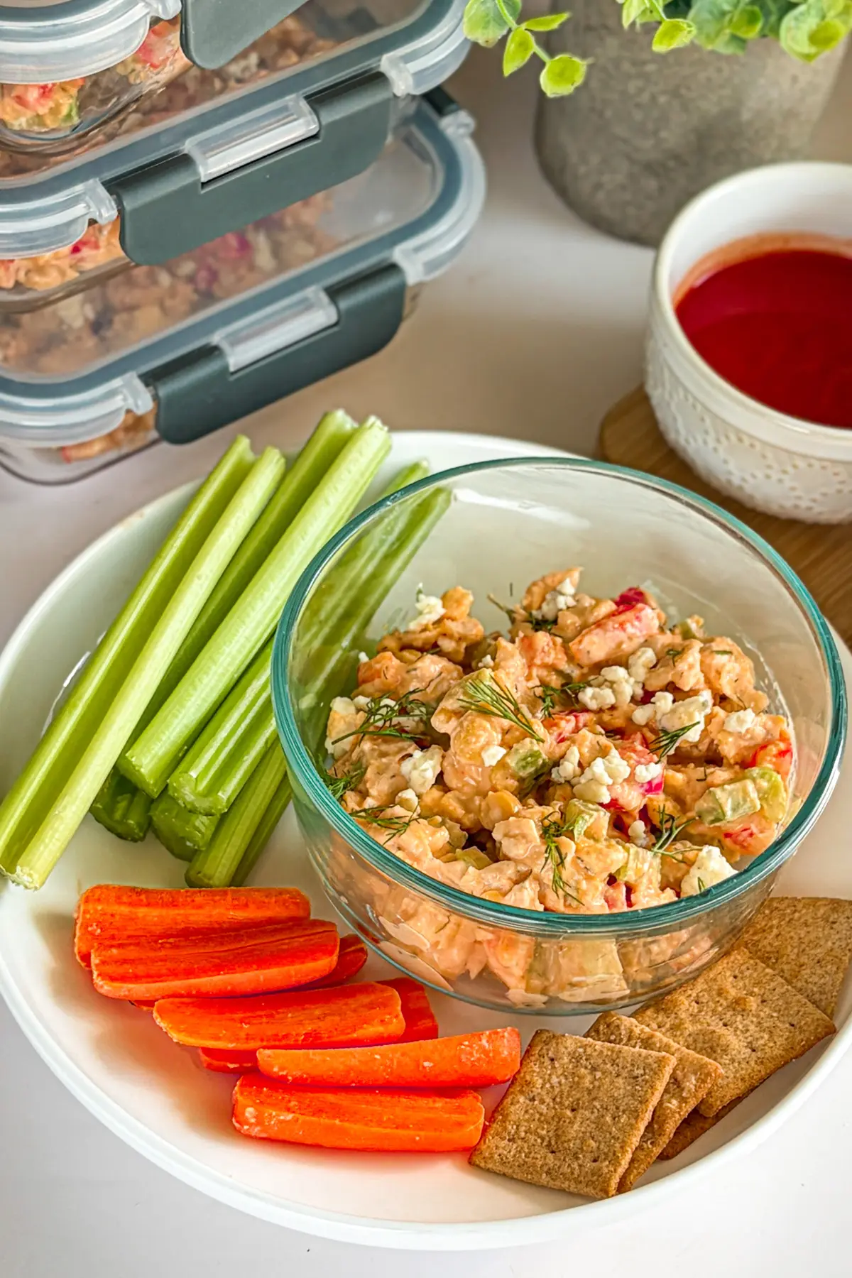 A loaded buffalo chicken dip snack plate with celery, carrots, and crackers on a table next to dip snack containers.