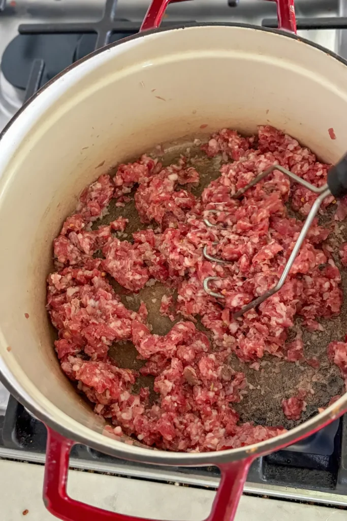 Raw Italian sausage being mashed with a potato masher in a Dutch oven.