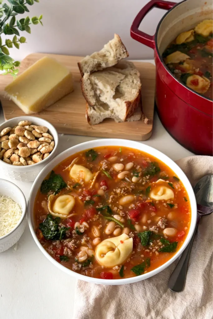 An overhead show of a bowl of soup on a table next to a big pot and a bowl of oyster crackers and a cutting board with crusty bread and a Parmesan rind.
