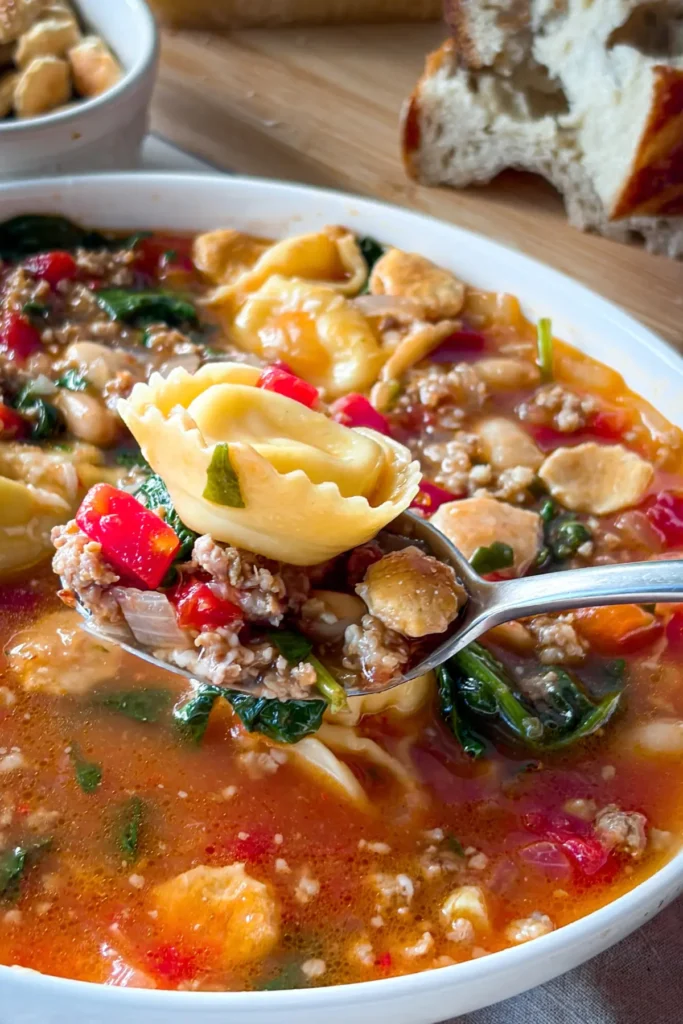 A close-up spoonful of the soup with a large tortellini on the spoon over a bowl of soup. 