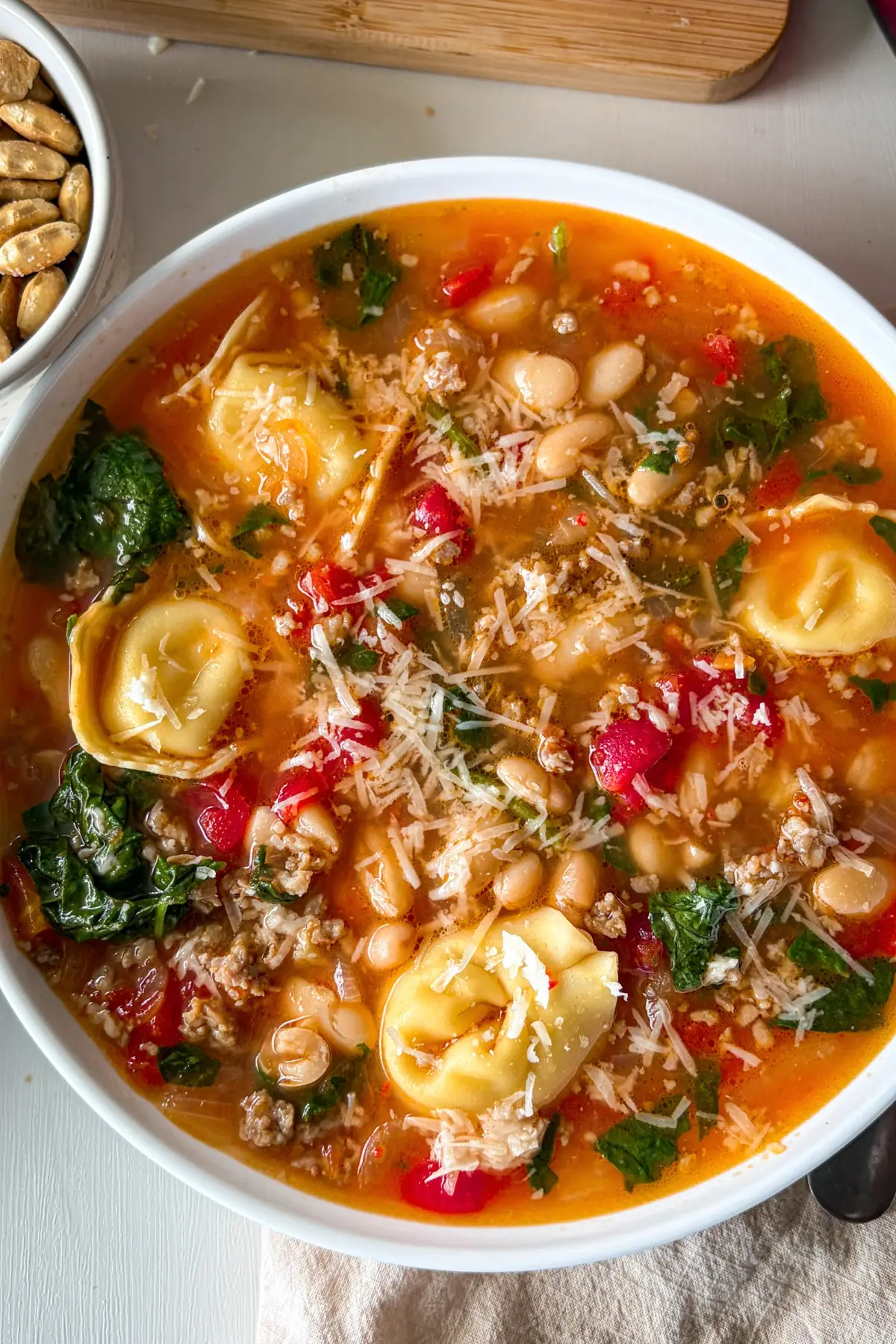A birds-eye view of a large bowl of Italian sausage tortellini soup with spinach on a table next to a bowl of oyster crackers and crusty bread.