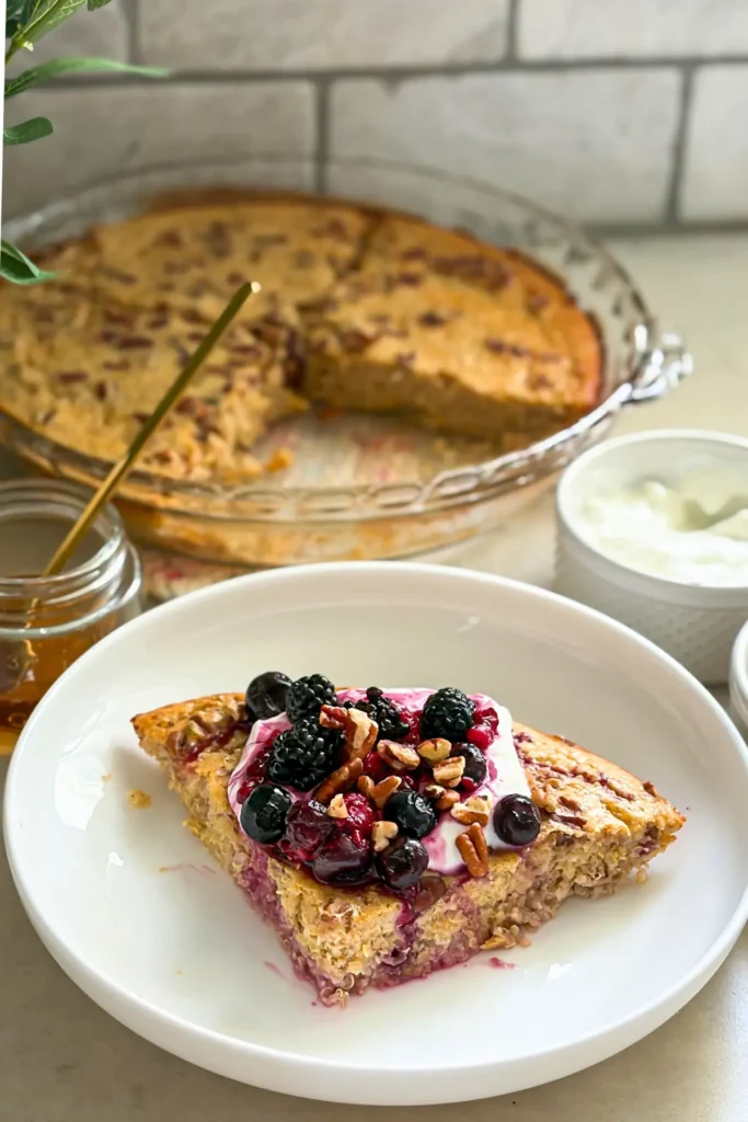A slice of the maple quinoa bake on a white plate topped with berries and yogurt next to a casserole dish with the remaining slices on a counter.