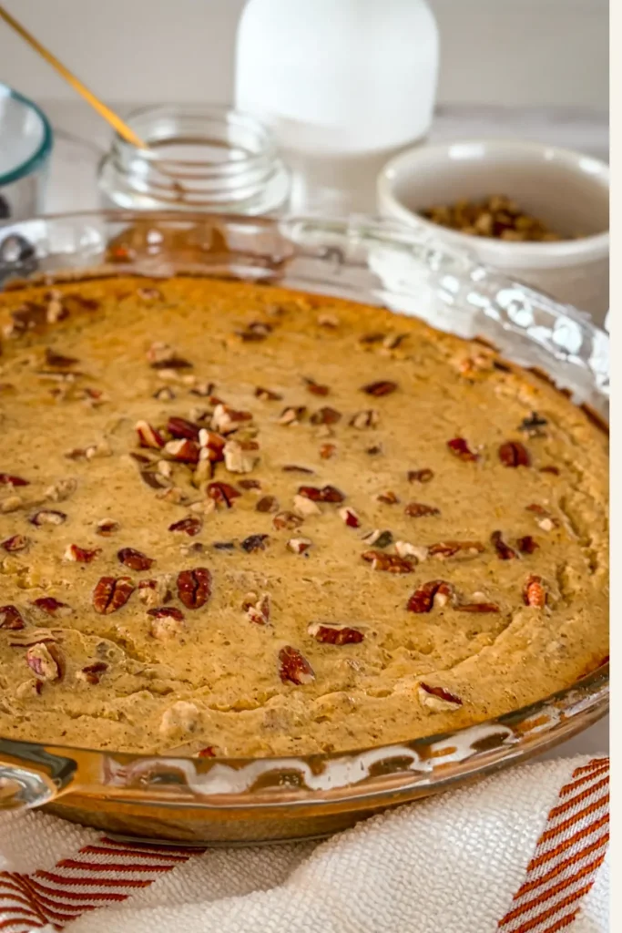 A maple pecan quinoa breakfast bake in a glass dish on a table next to a bowl of maple syrup, a bowl of chopped pecans and a spatula.